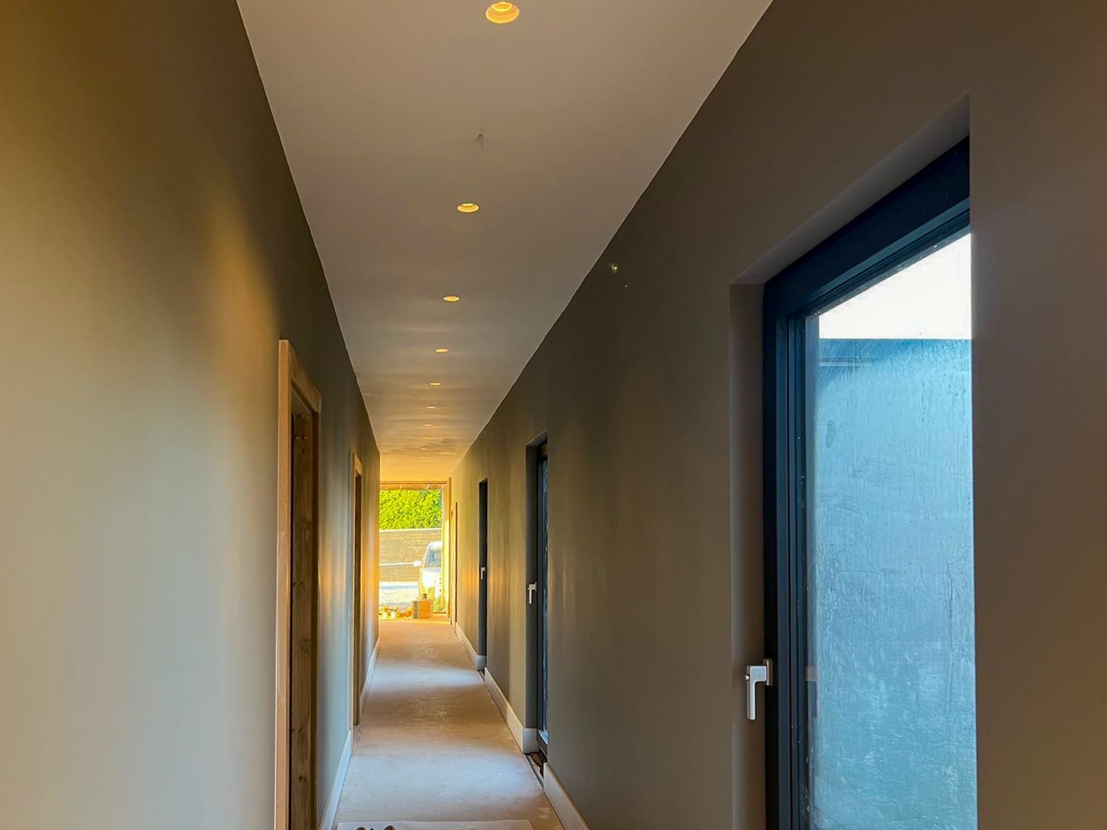 Long hallway with recessed ceiling lights, timber doors and a large side window.