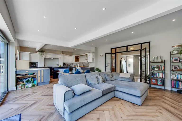 Open-plan lounge and kitchen with a grey corner sofa, herringbone flooring and black-framed doors